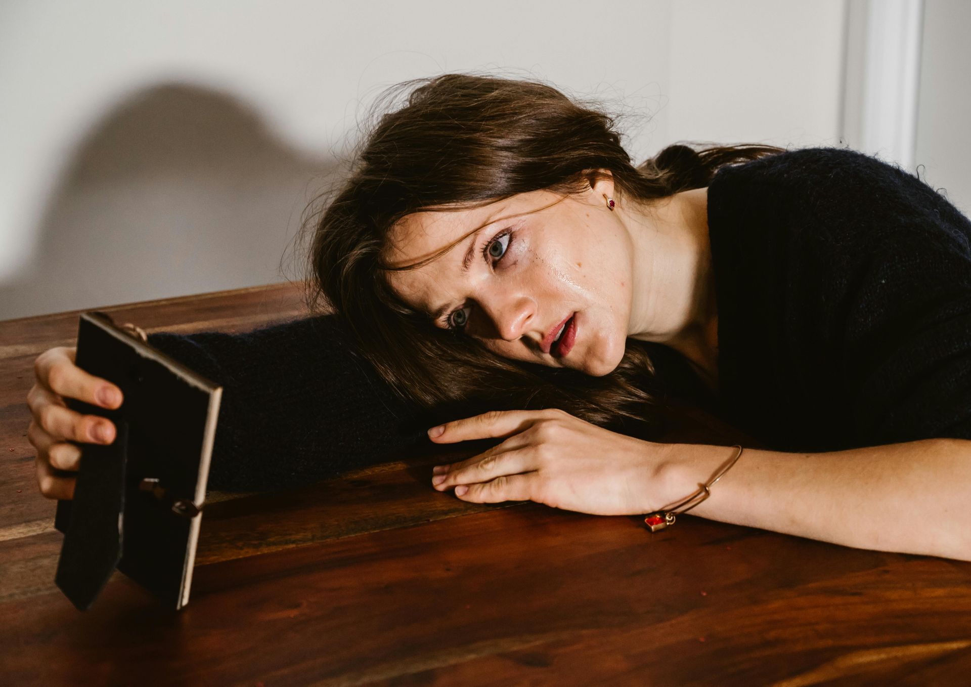 A person in a black sweater rests their head on a wooden table, sadly looking at a small, framed photo.