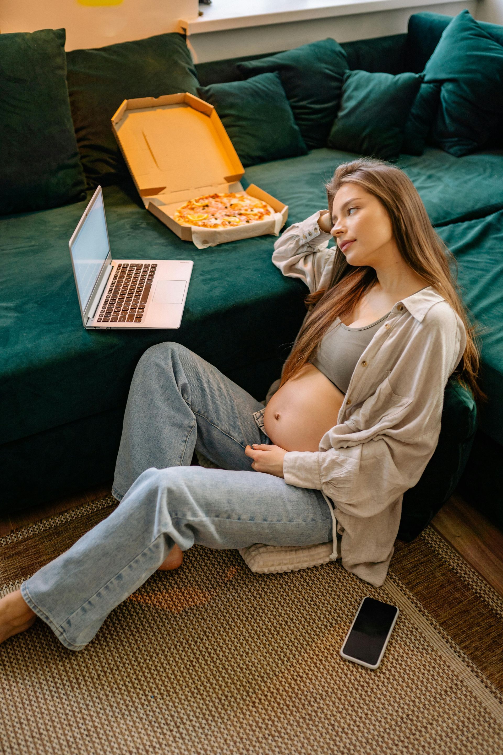 A pregnant person sits on a rug by a green sofa with a laptop and a box of pizza nearby.