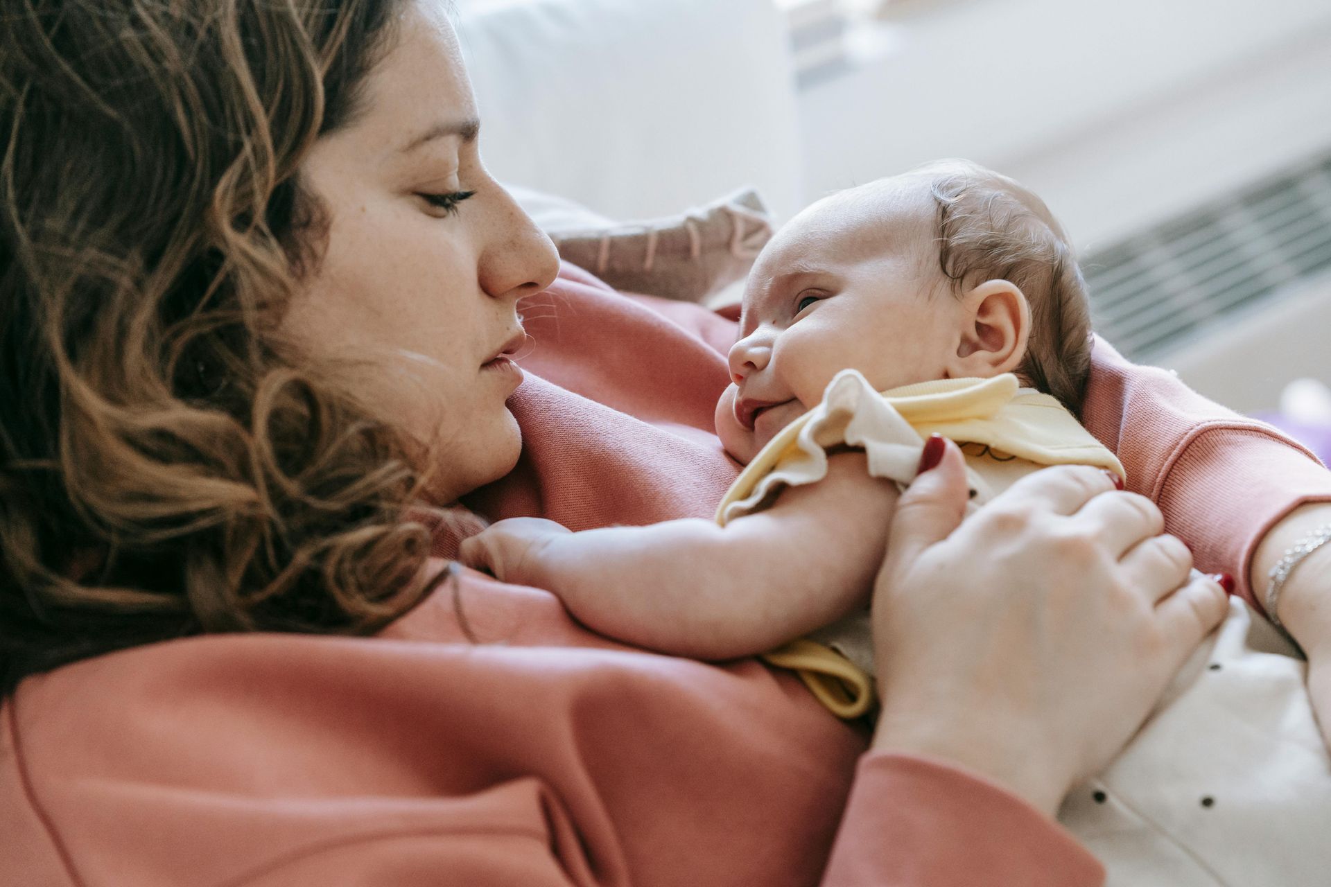 A person in a pink sweater holds a baby wearing a yellow shirt, looking at them with a gentle expression.