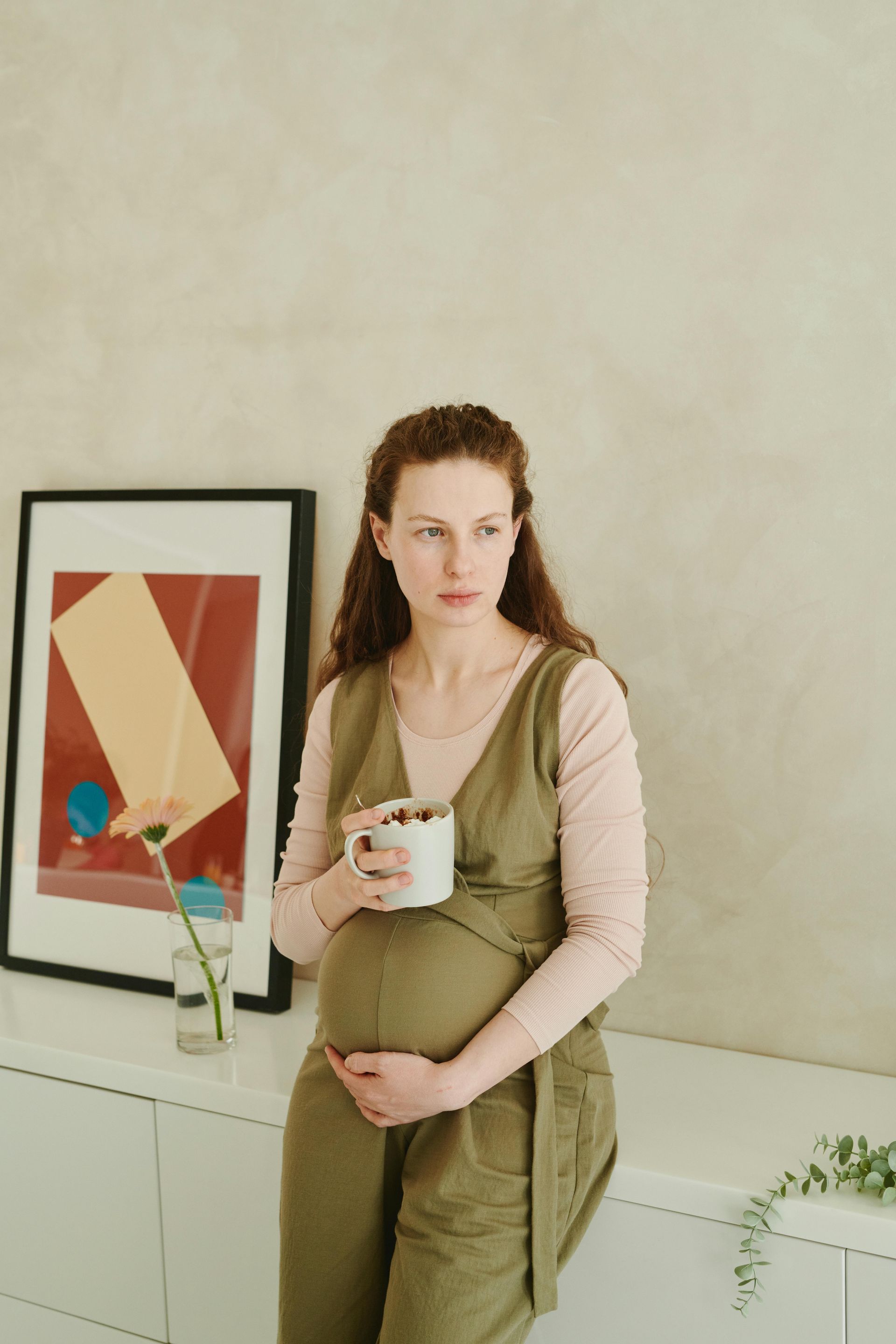 A pregnant person in olive green overalls and a pink long-sleeved shirt holds a mug while leaning against a white counter.