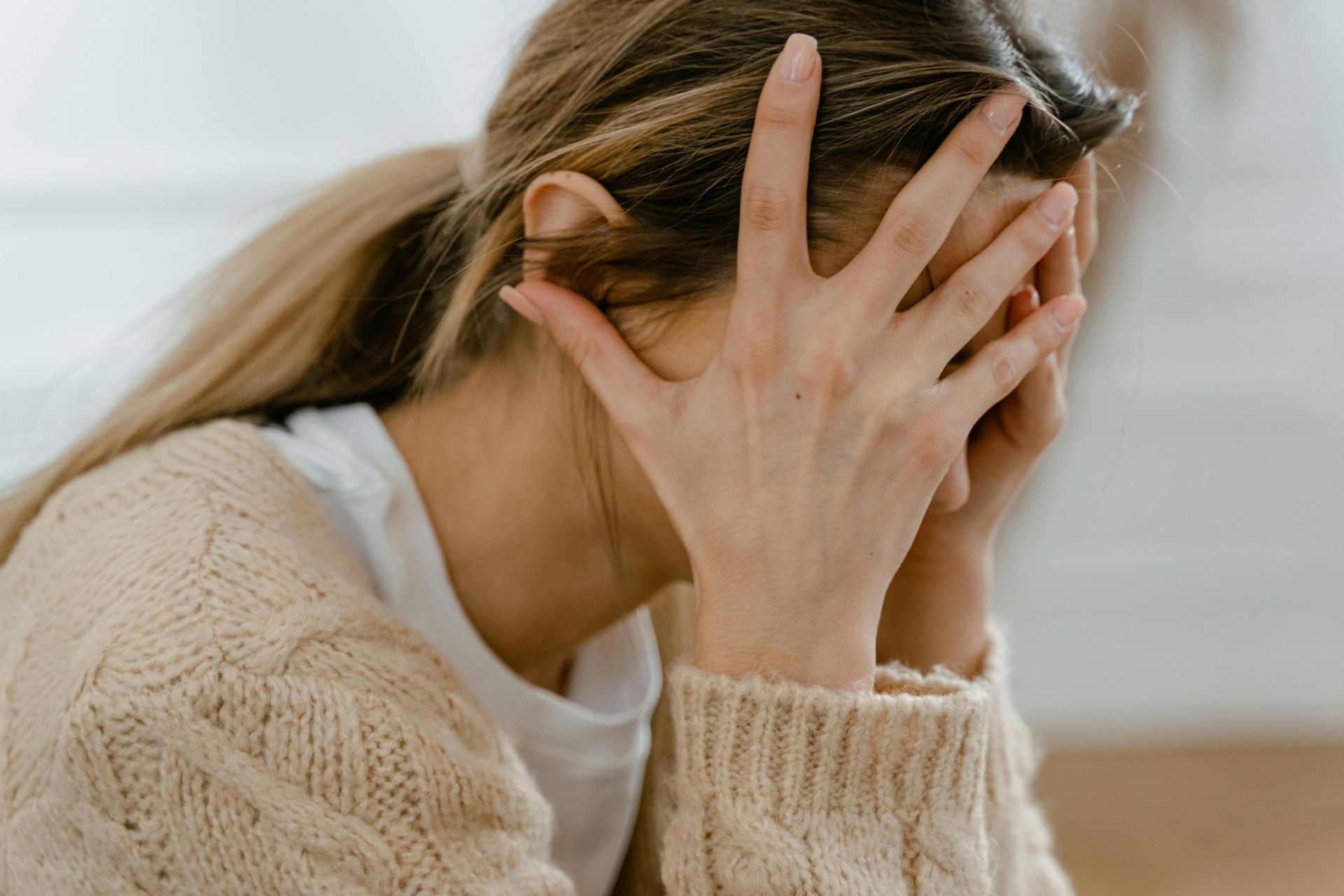 A person with long blonde hair wearing a beige knit sweater, holding their face in their hands in a gesture of distress.
