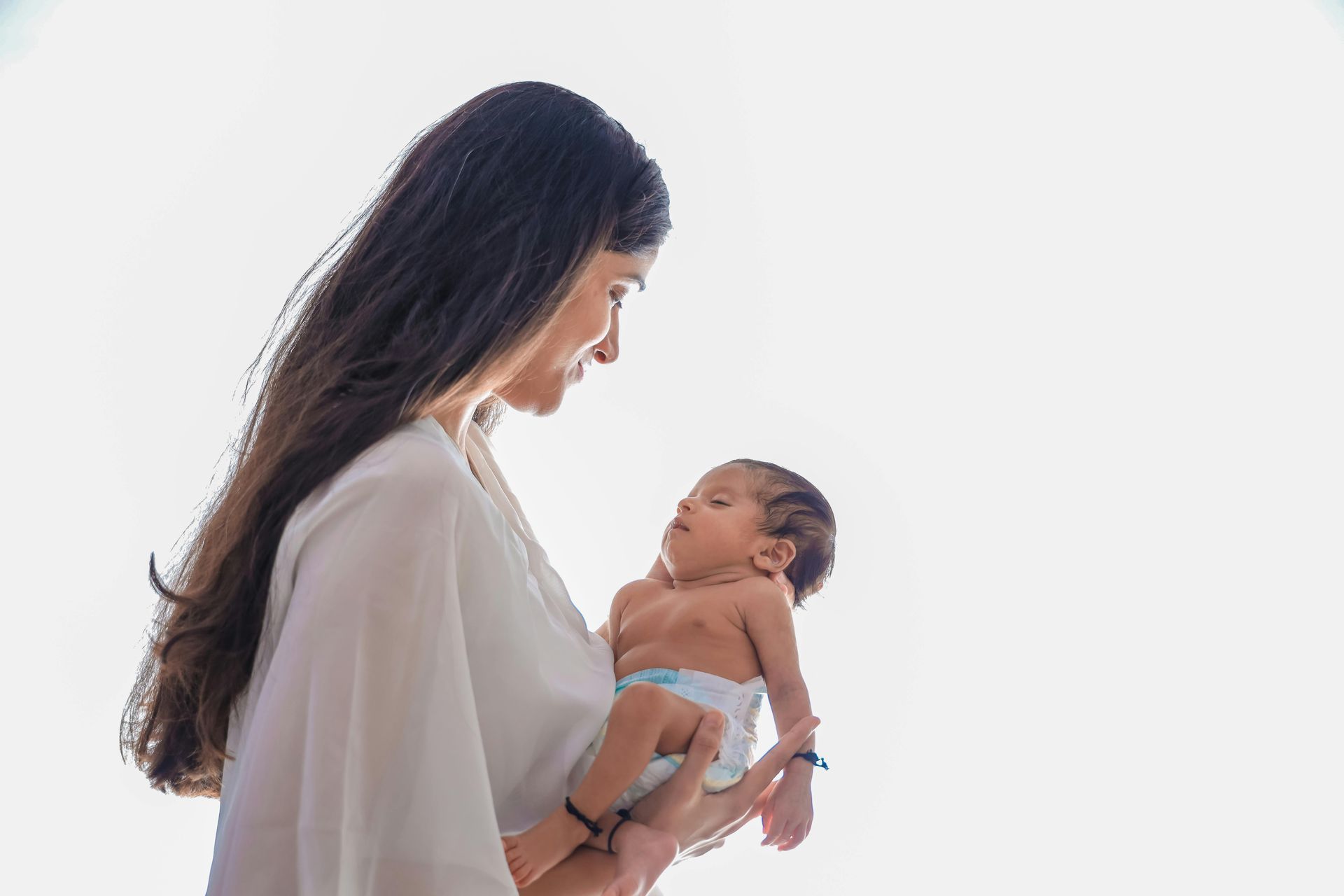 A woman with long dark hair holds a baby against a plain white background.
