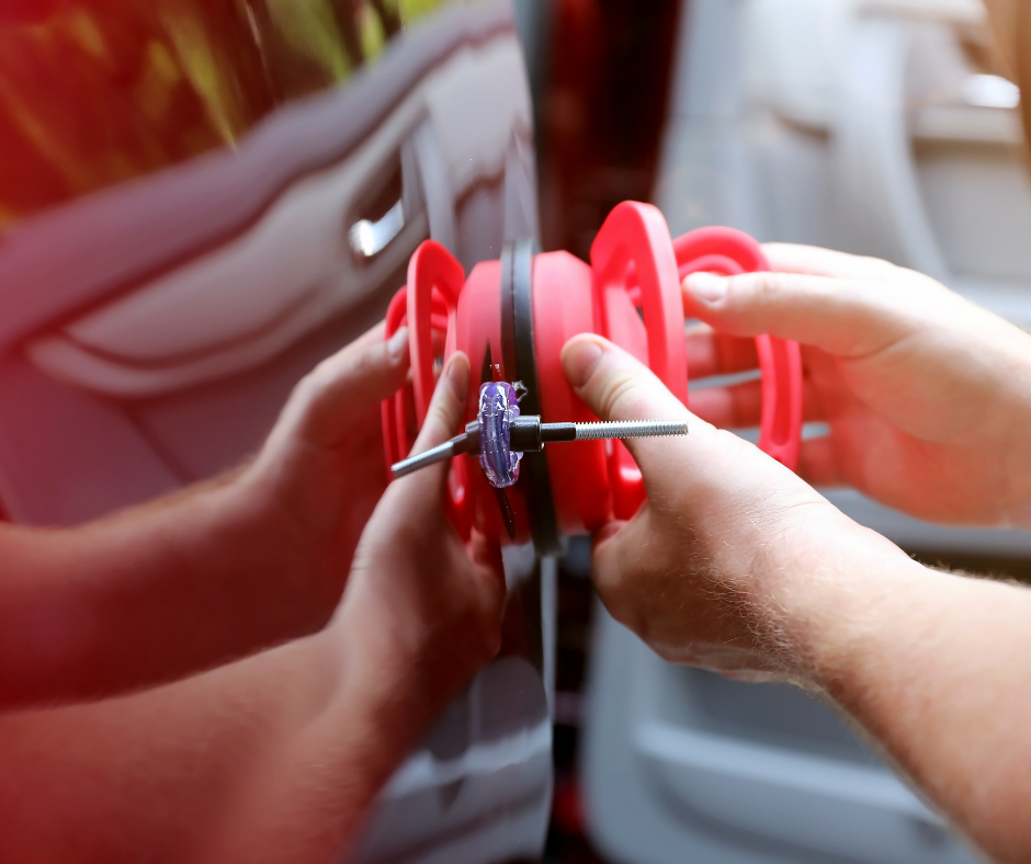 Hands holding a red wheeled toy or tool against a car door, with a metal shaft centered between the wheels.