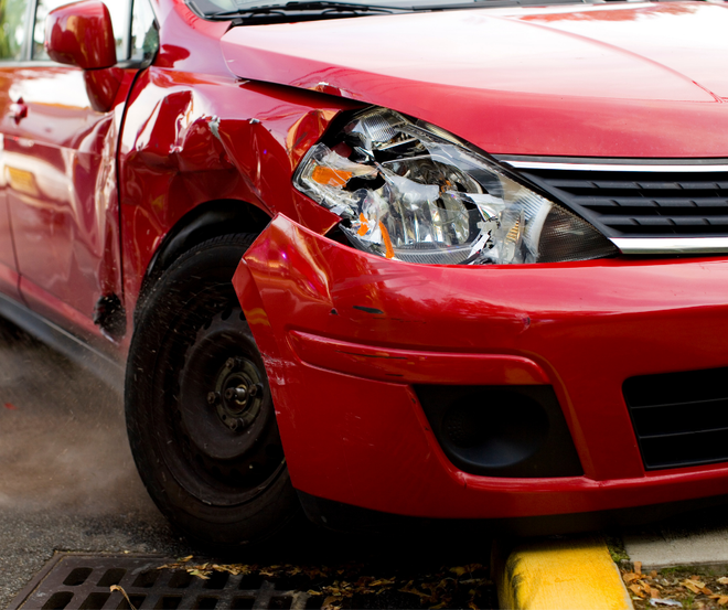 Red car with a damaged front fender and headlight, parked near a curb with smoke rising from the wheel area.