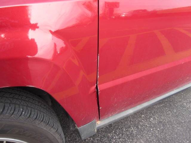Close-up of a red car’s side panel with a dent and scrape near the rear wheel.