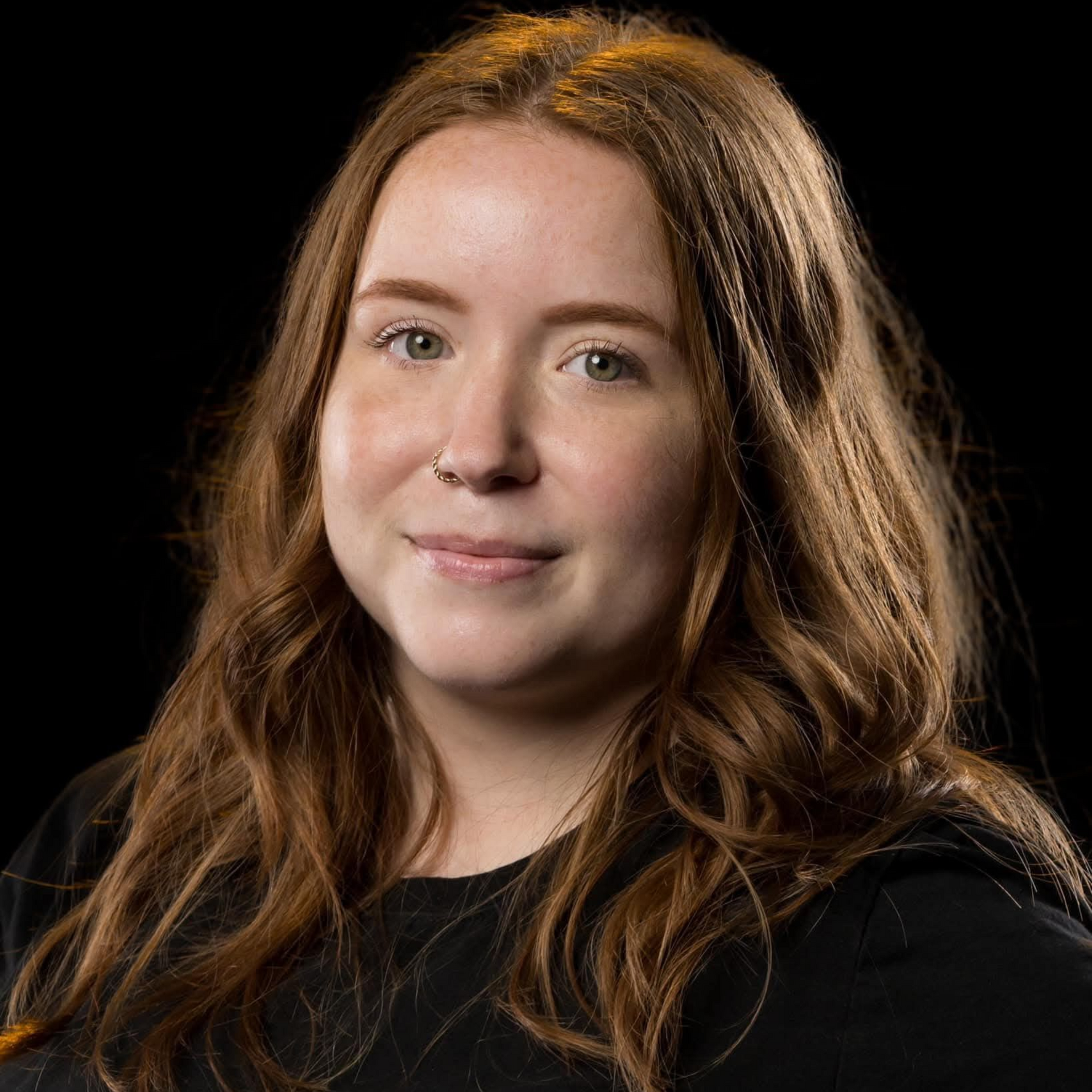 Portrait of a person with wavy brown hair on a black background, wearing a black top, slight smile