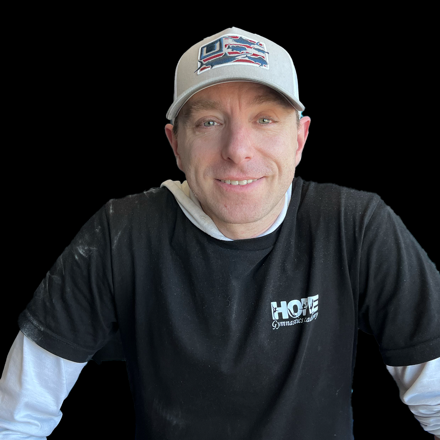 Smiling man in a gray cap and black shirt over a white long-sleeve shirt, seated against a black background