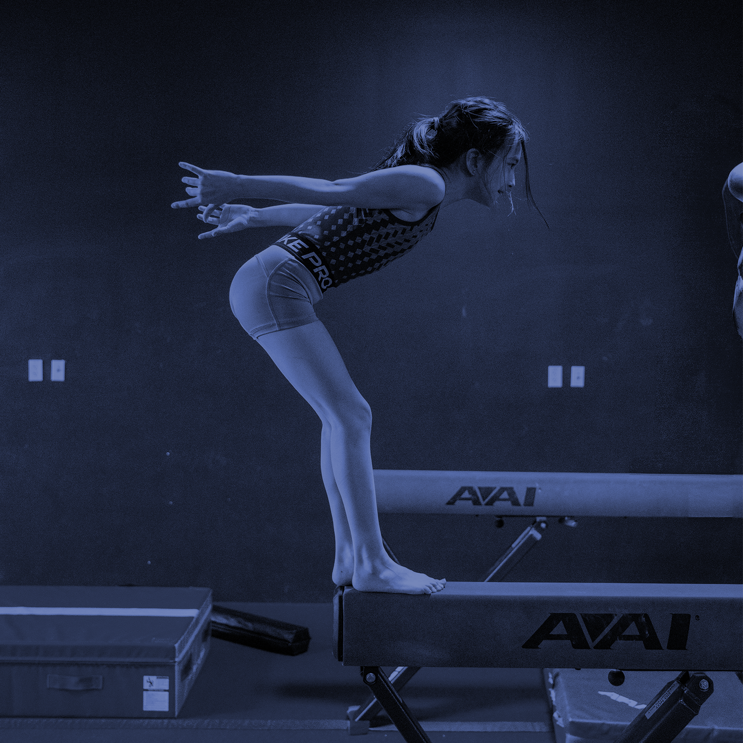 Gymnast balancing on a beam in a dark, blue-lit gym, arms extended behind her.