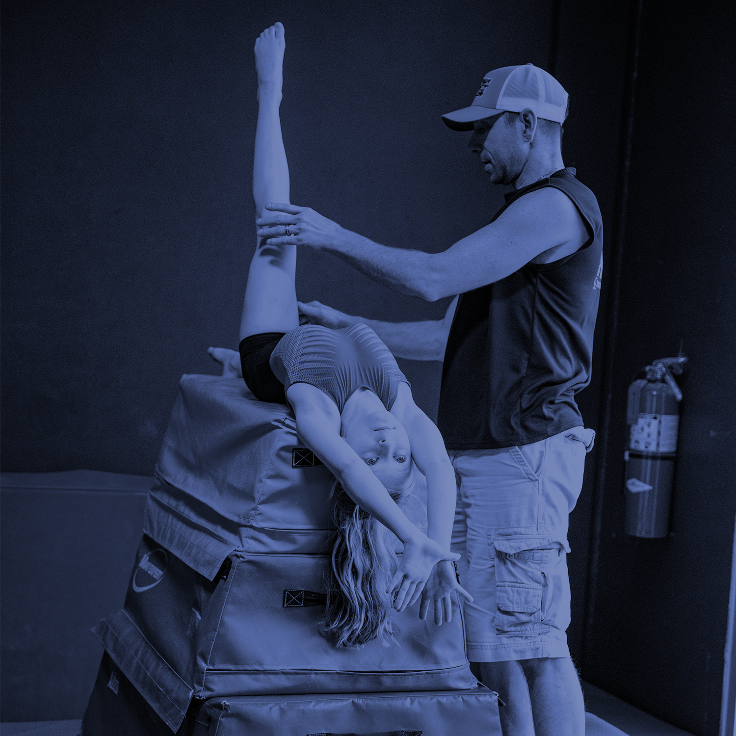 Martial arts instructor assists a student in a stretching pose in a gym, with a fire extinguisher in the background