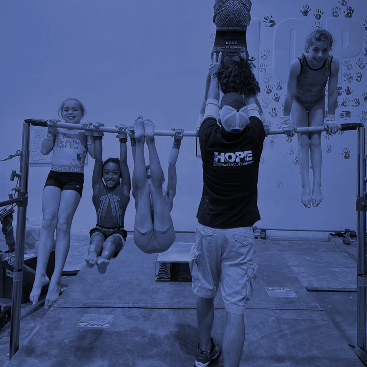 Children doing gymnastics on parallel bars in a gym, with a coach spotting them upside down.