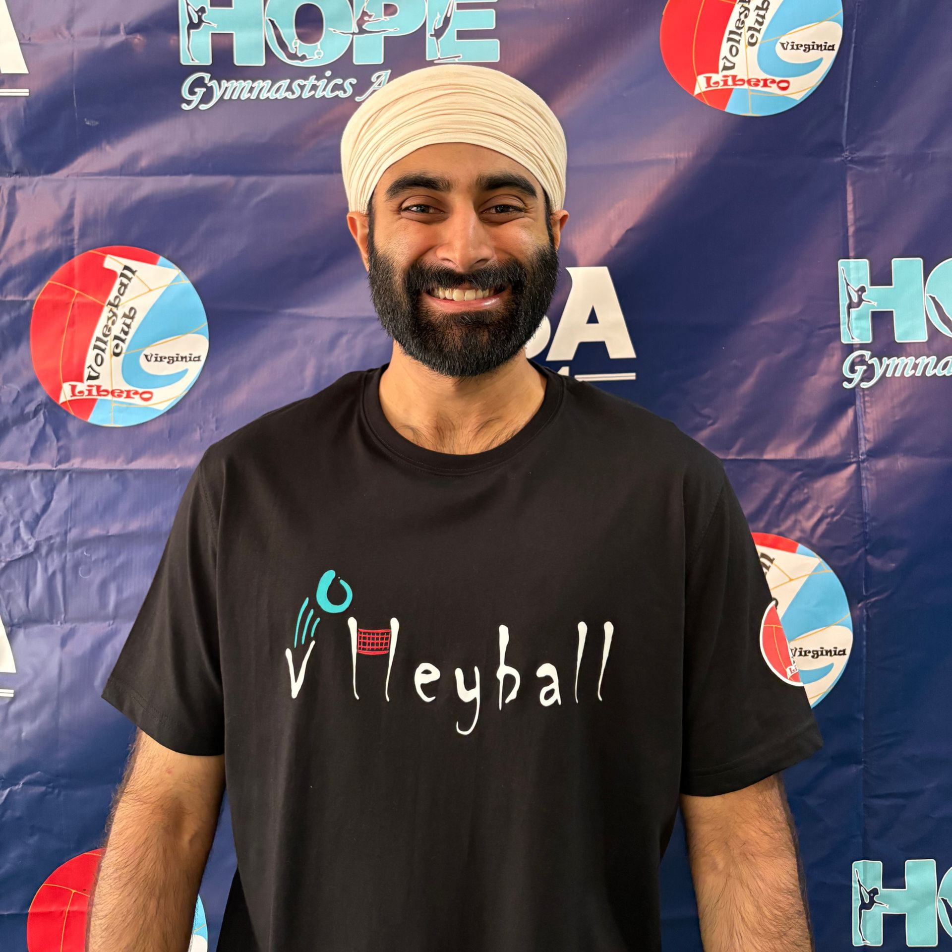 Smiling person in a black T-shirt and pink turban, arms crossed against a black background.