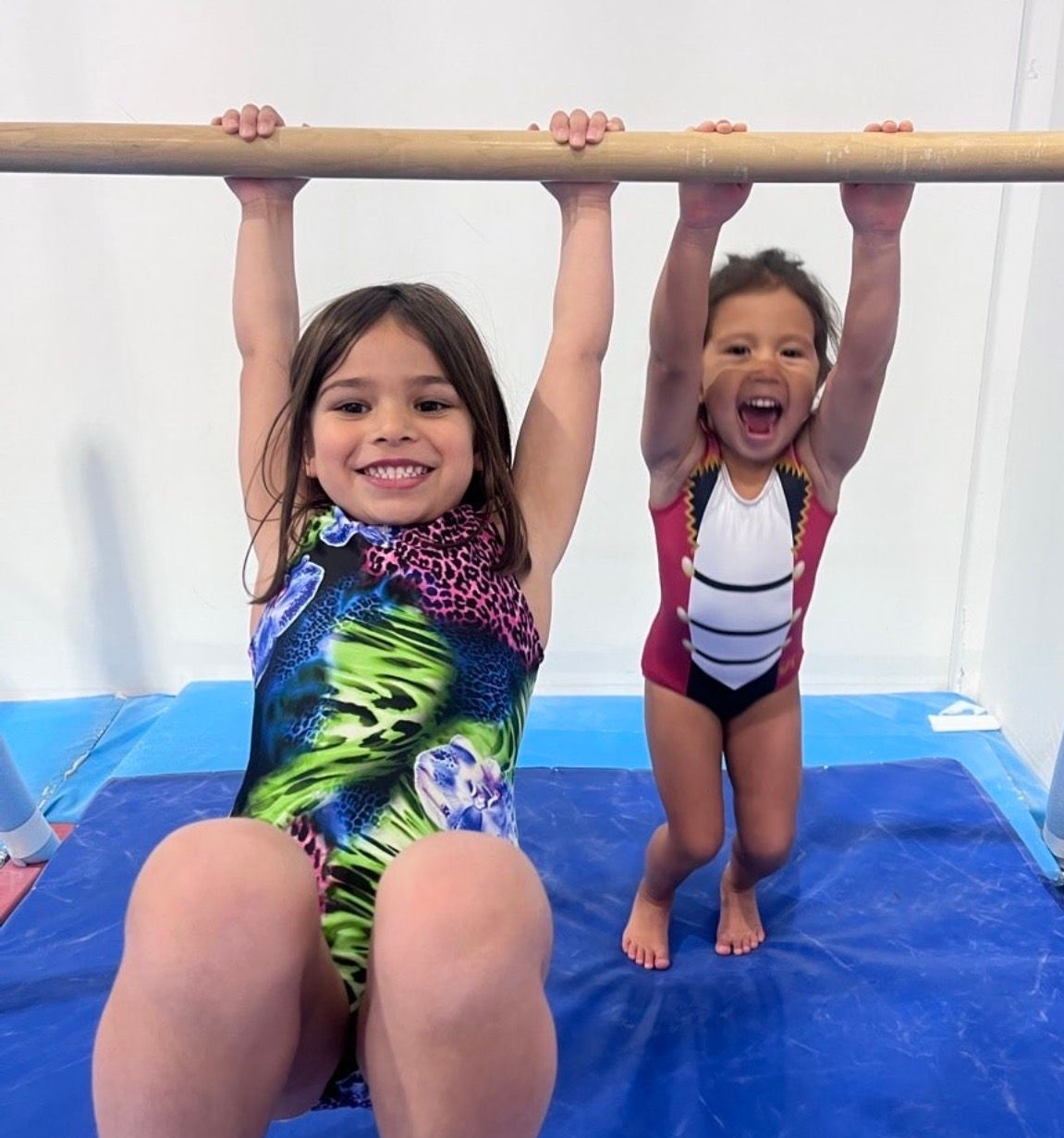 Two children hanging from a wooden bar on blue mats, smiling in a gym or studio.