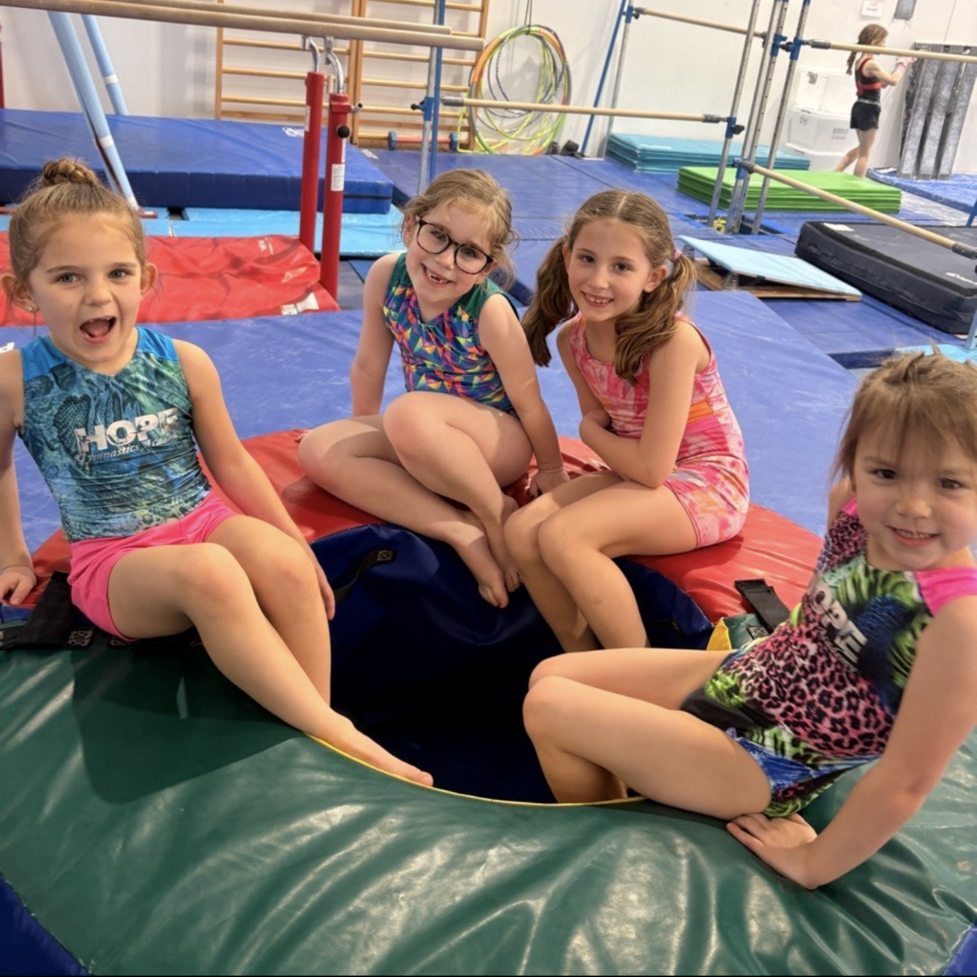 Four children sit on colorful gym mats, smiling in a gymnastics studio with equipment in the background.