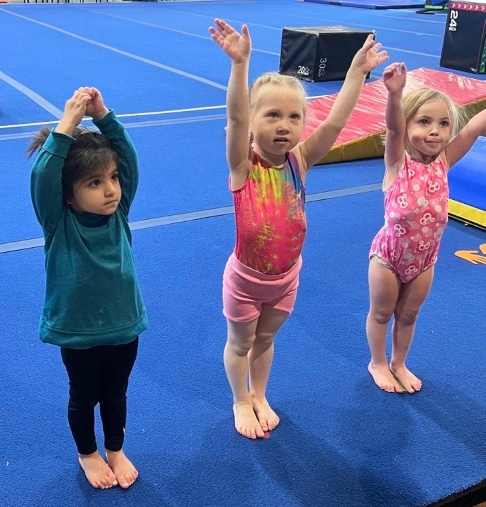Three young gymnasts standing on a blue mat with arms raised during practice