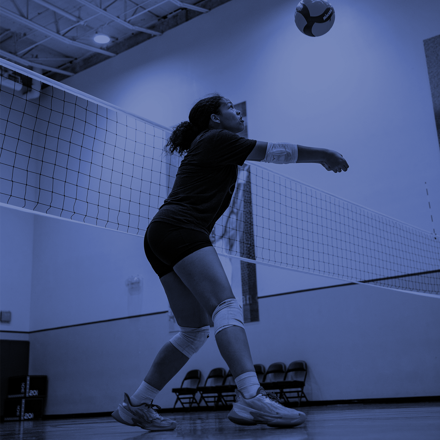 Indoor volleyball player in mid-serve, reaching toward the ball near the net in a gym.
