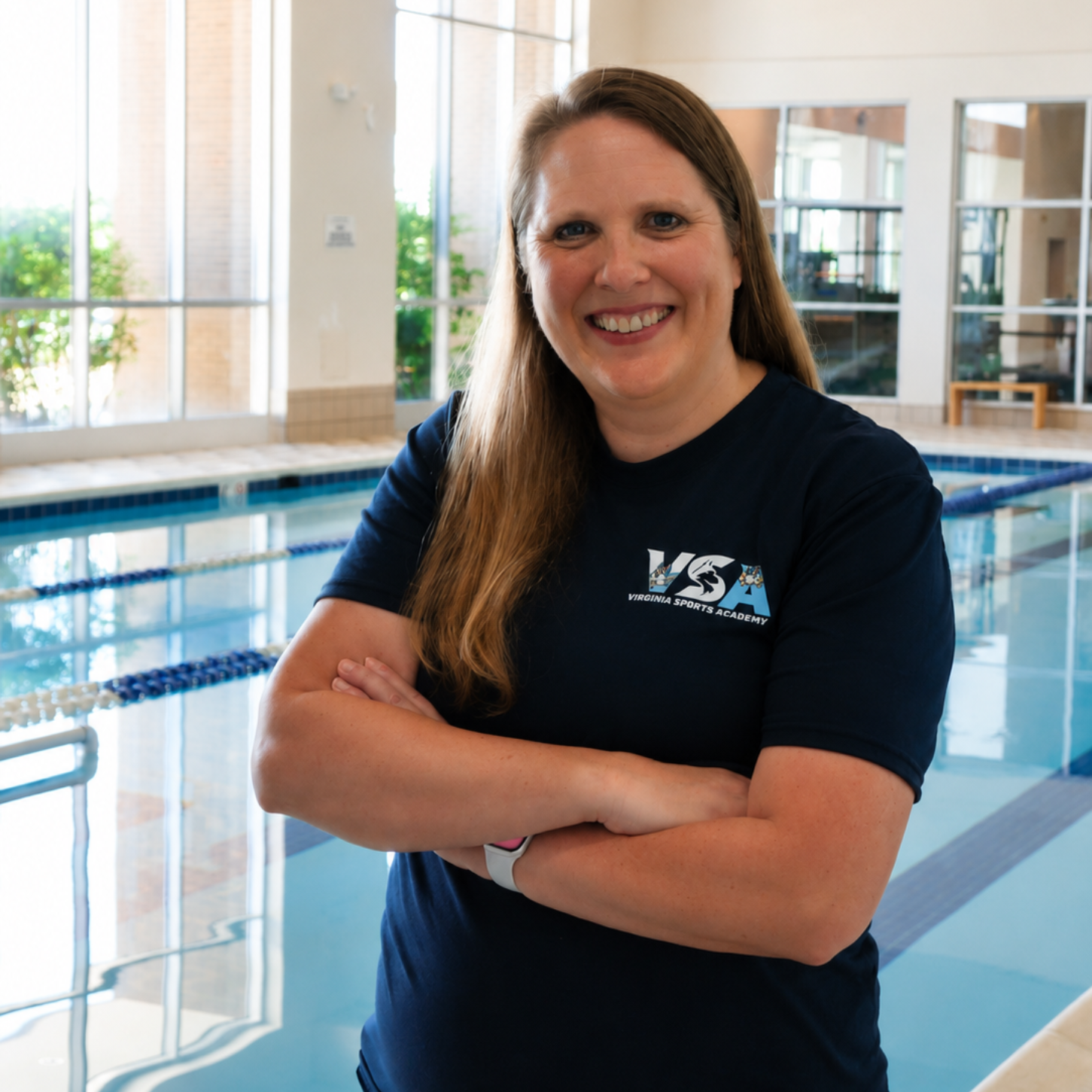 Woman in navy shirt smiling with arms crossed by an indoor swimming pool