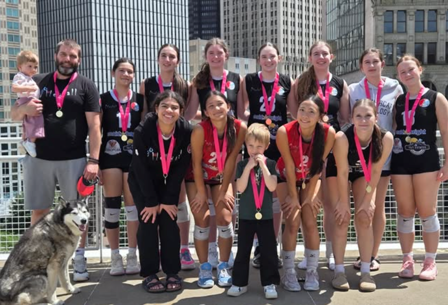 Volleyball team posing with medals and trophy outdoors, with a dog in front of the group.