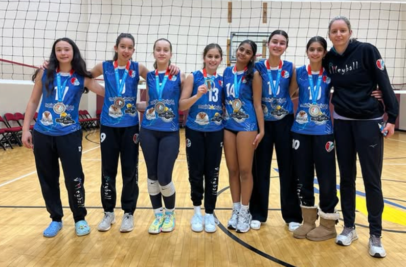 Volleyball team posing on an indoor court with medals and blue jerseys, standing beside their coach
