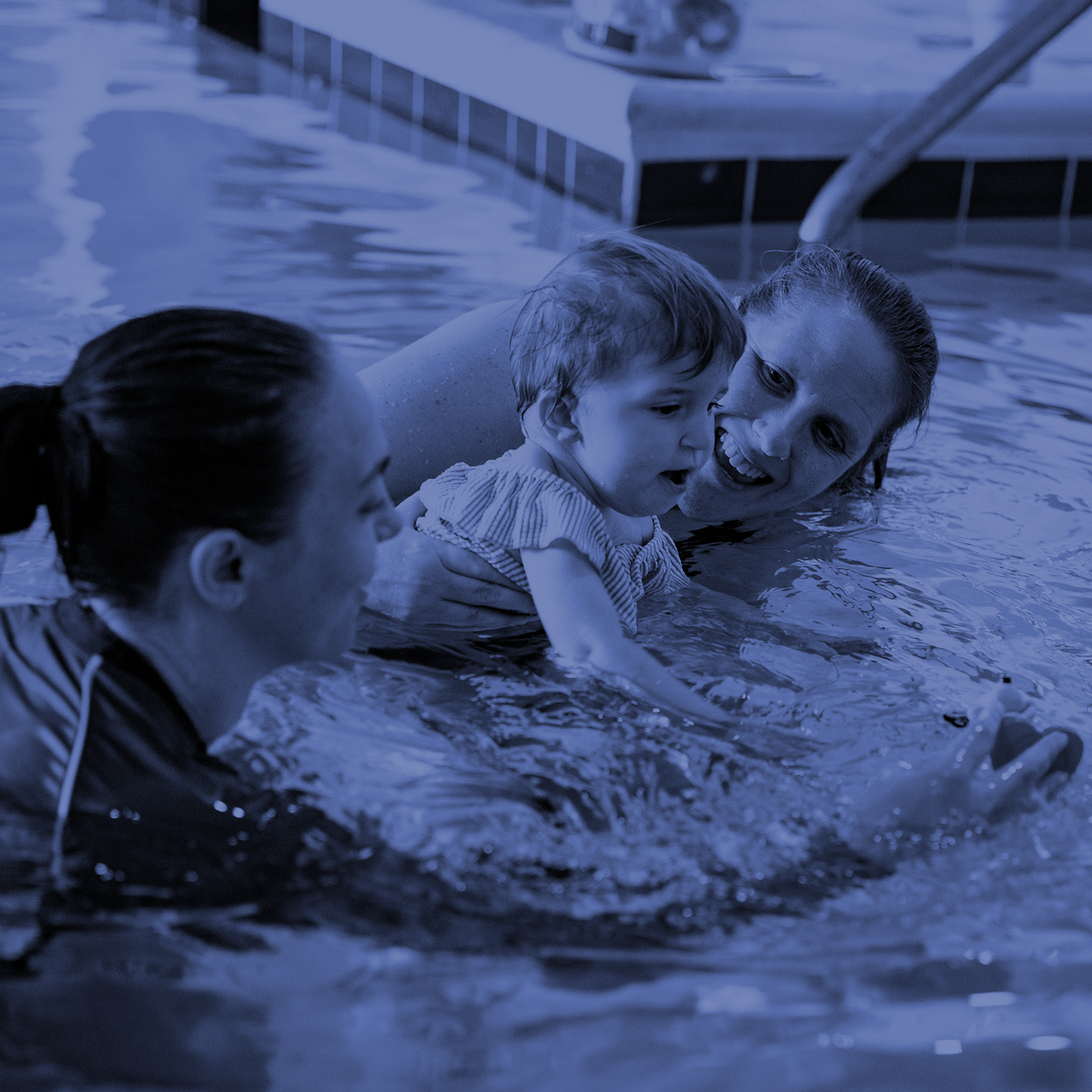 Three people in a swimming pool helping a toddler float and smile near the water surface.