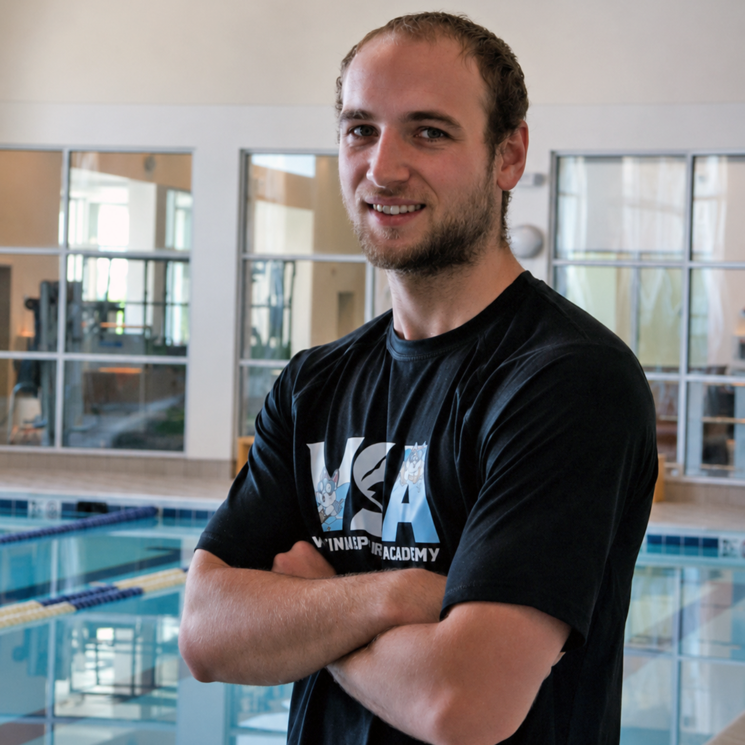 Man with crossed arms standing by an indoor swimming pool, wearing a black T-shirt and smiling.