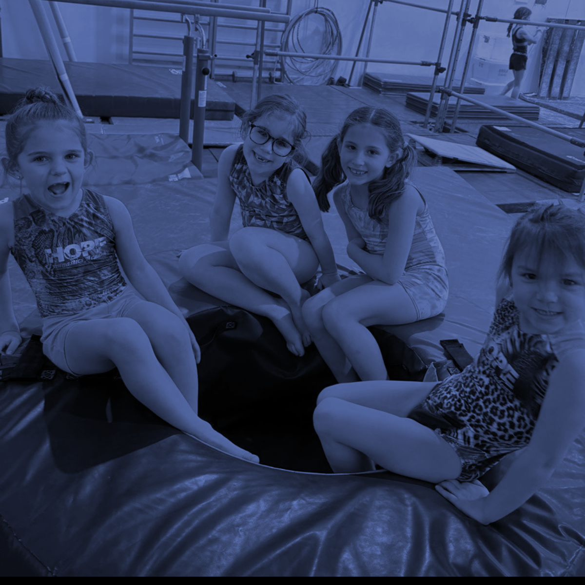 Four children sitting on a padded mat in a gymnastics gym, smiling at the camera.
