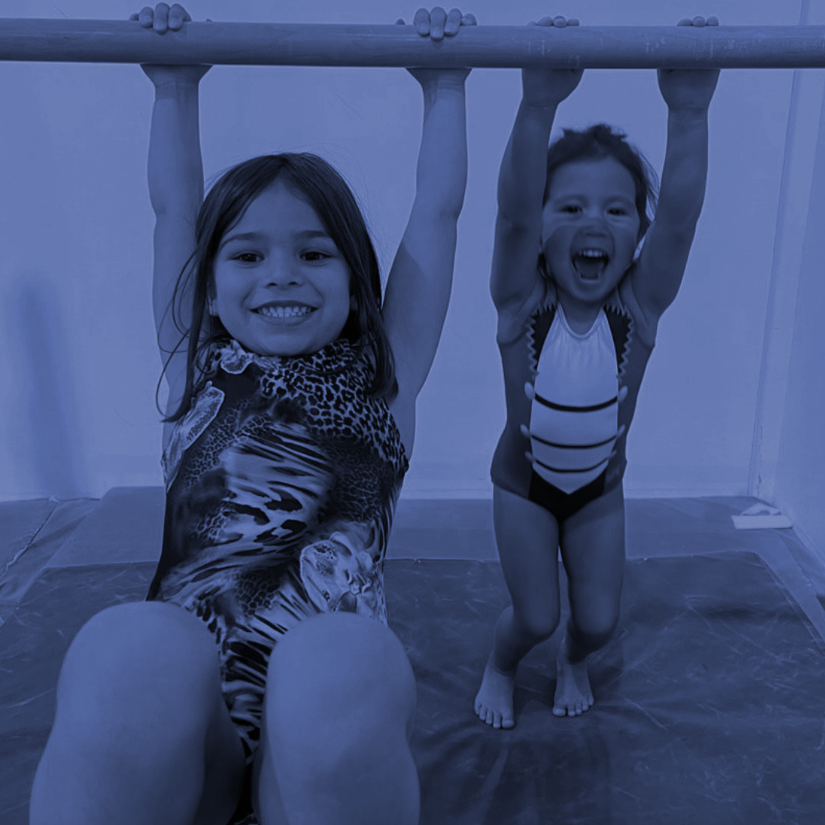 Two children hanging from a horizontal bar, smiling indoors on mats.