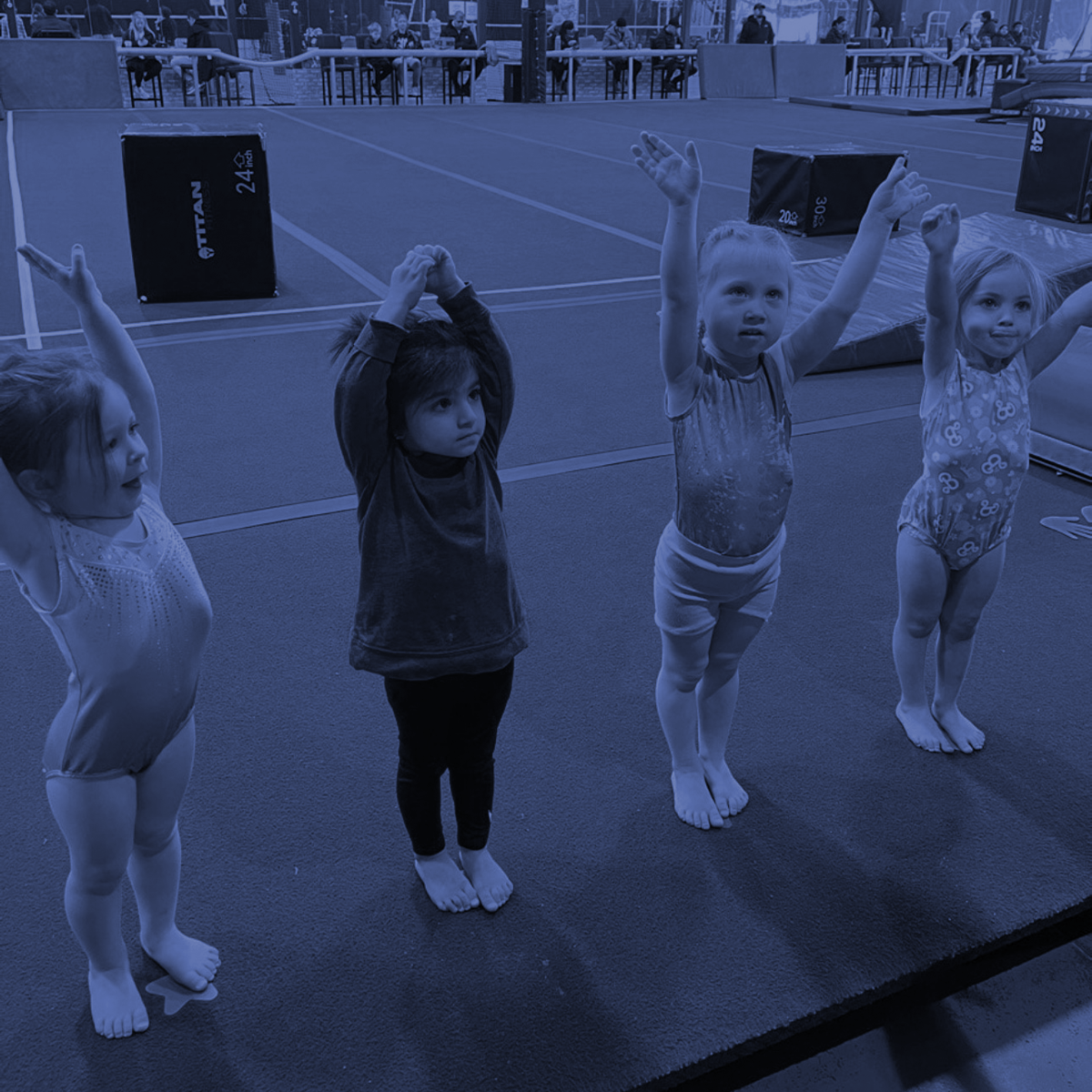 Four toddlers in leotards stand on a mat with arms raised during an outdoor dance class.