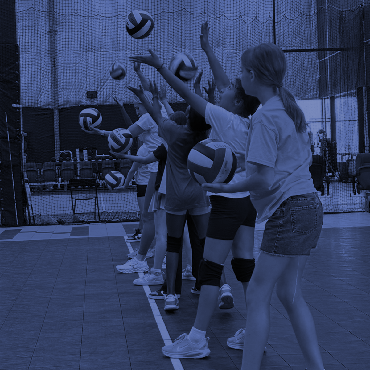 Volleyball players practice setting and spiking balls in an indoor gym, shown in blue tint.