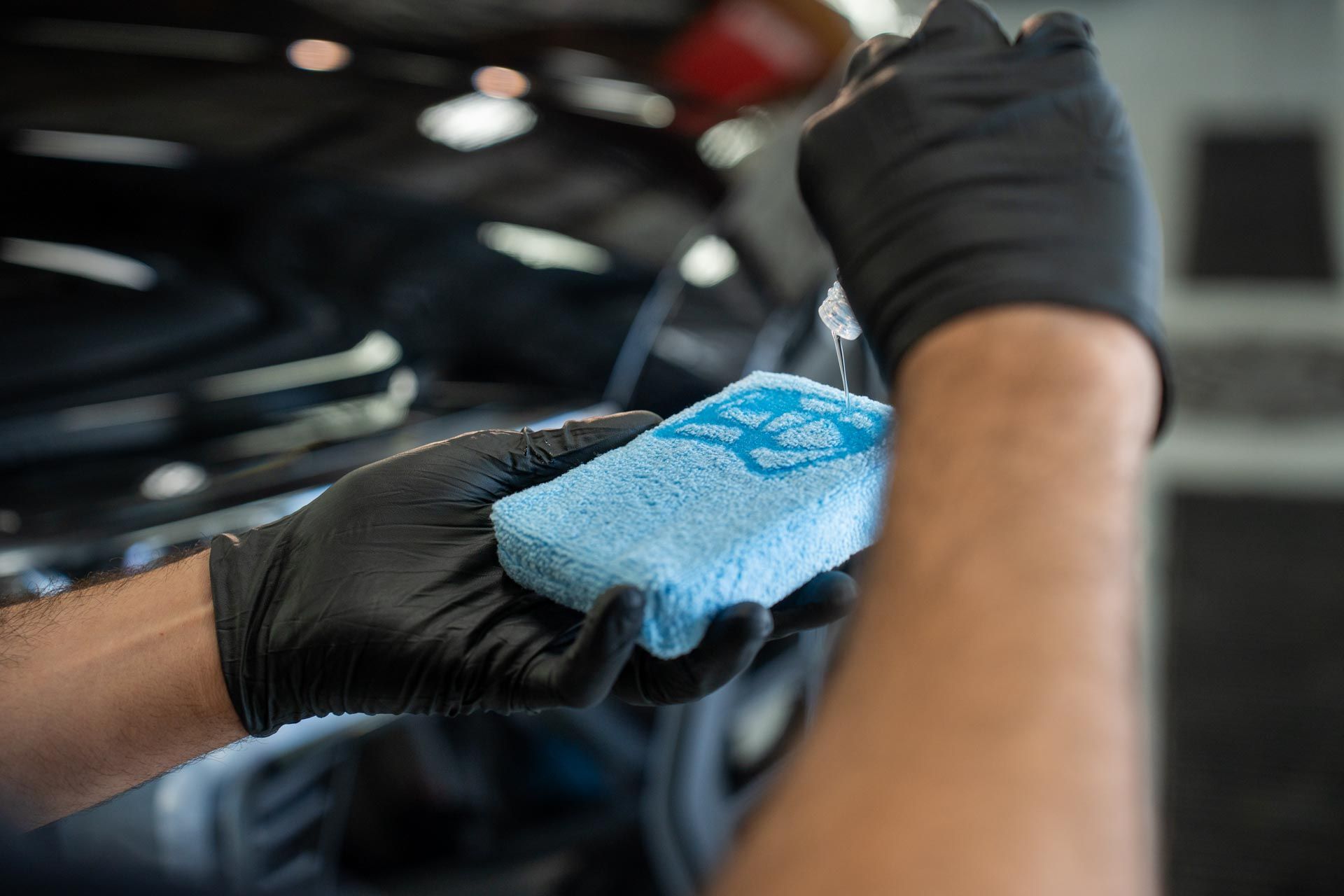 Person wearing black gloves spraying liquid onto a blue sponge, likely cleaning a car.
