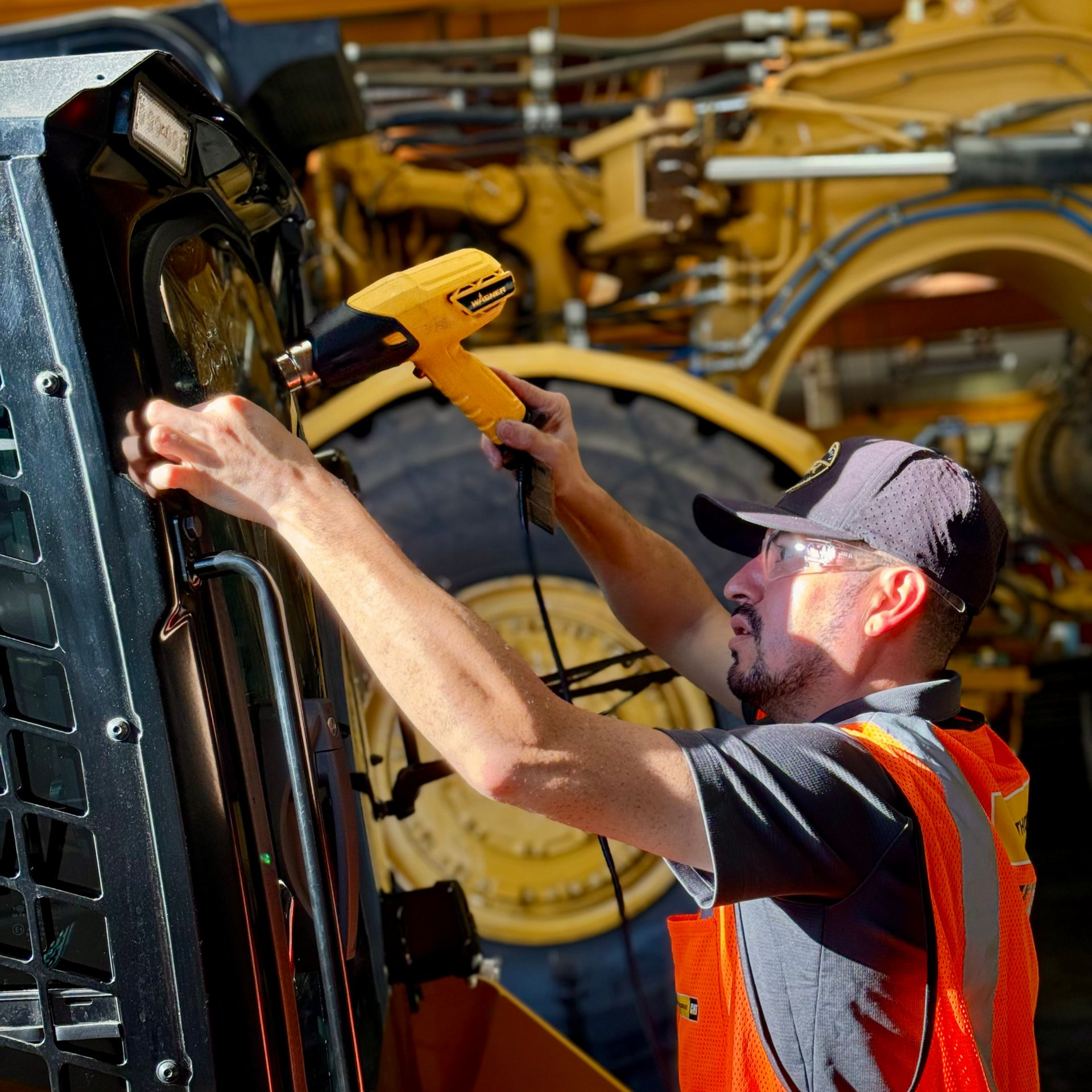 Mechanic using a heat gun on heavy machinery, wearing safety vest and cap. Yellow and black colors dominate.