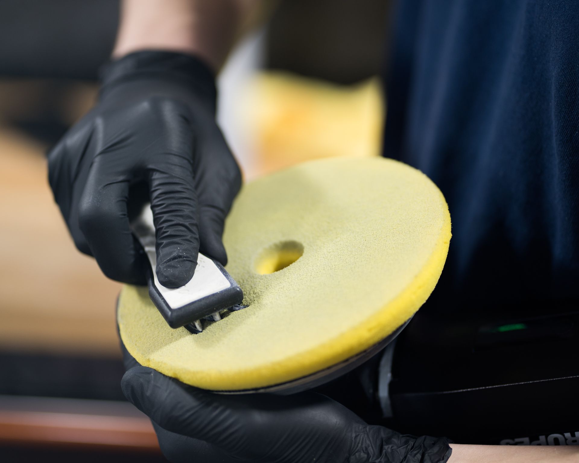 A person wearing black gloves cleans a yellow buffing pad with a brush.