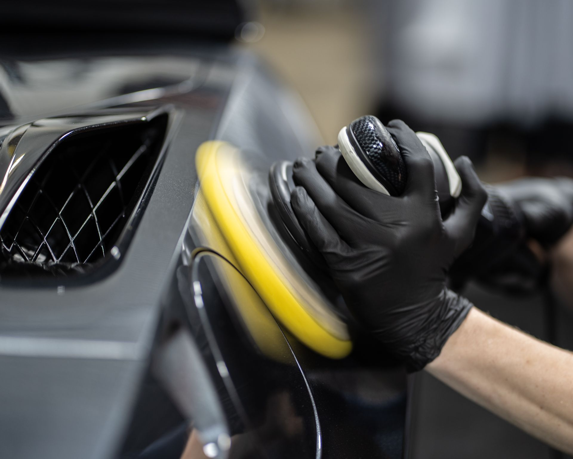 Person polishing a gray car with a yellow buffing pad and black gloves.