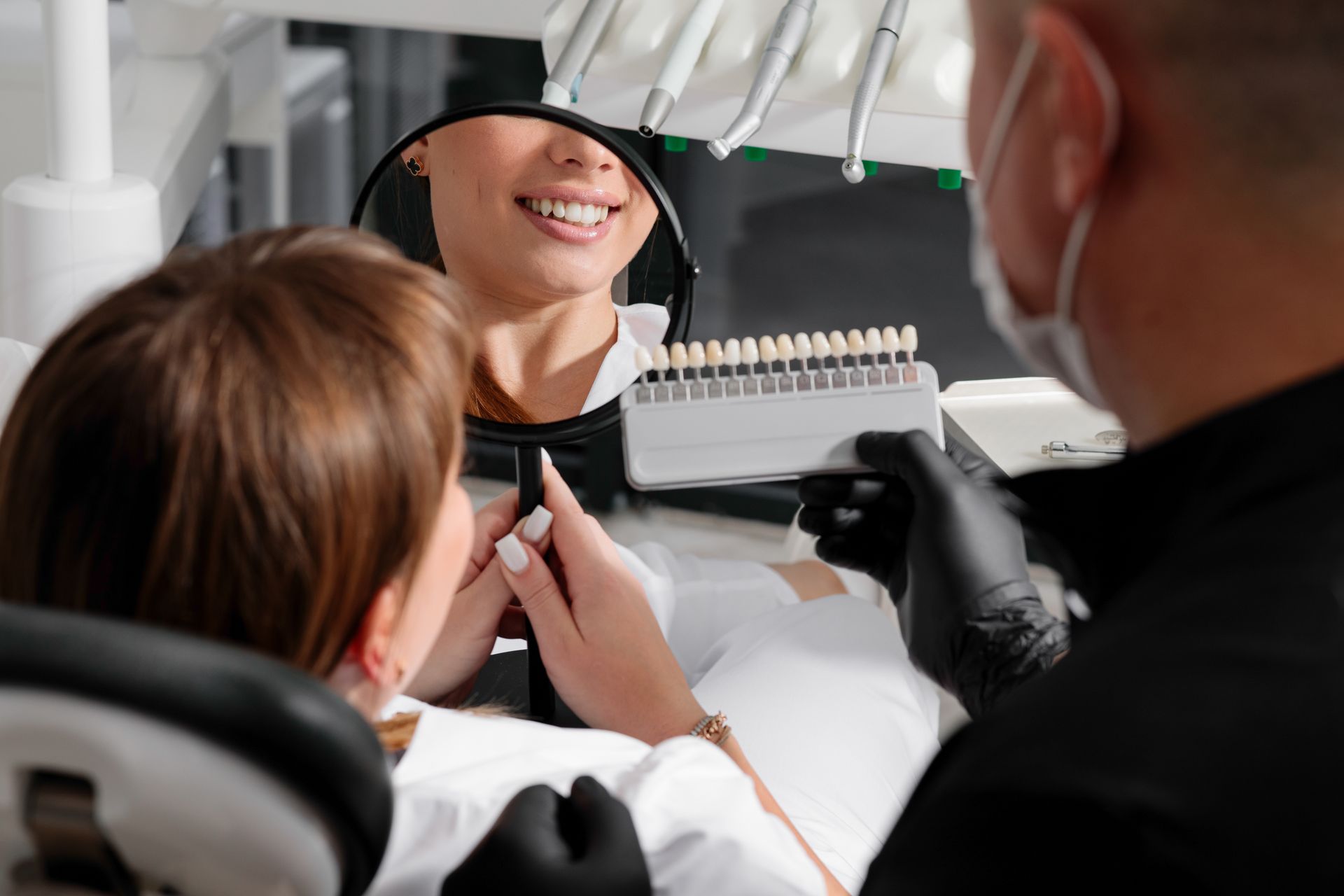 A smiling woman holds a mirror while a dentist shows her a tooth shade guide.