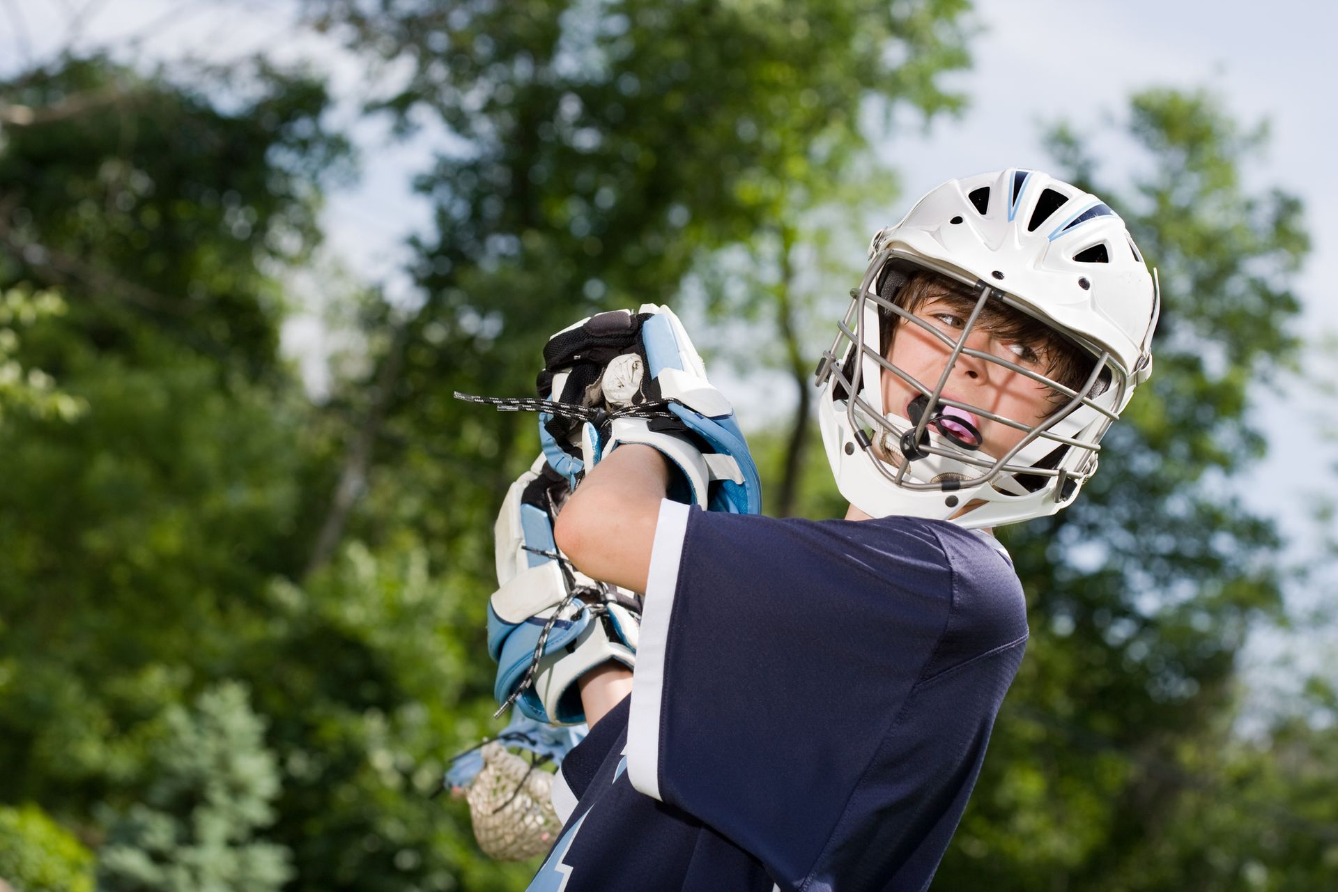 Youth lacrosse player in navy jersey and white helmet holding a stick outdoors with trees behind.