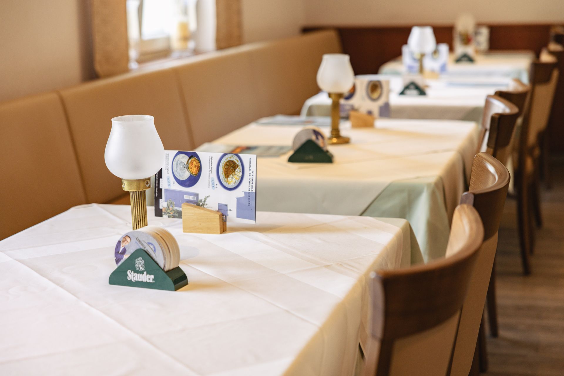 Restauranttische mit Lampen, Speisekarten und Untersetzern. Hellbraune Sitzecken und Holzstühle.