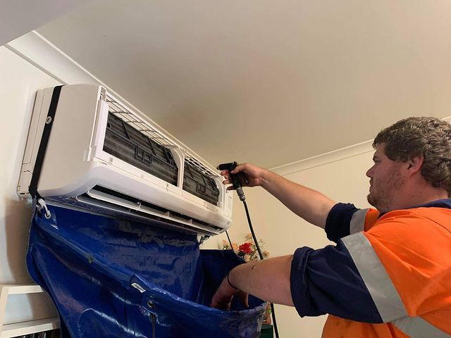 A Man Is Cleaning an Air Conditioner with A Hose — Paton's Electrical Services in Hervey Bay, QLD