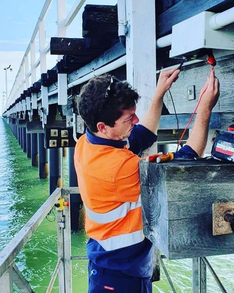 A Man in An Orange Shirt Is Working on A Bridge — Paton's Electrical Services in Urraween, QLD