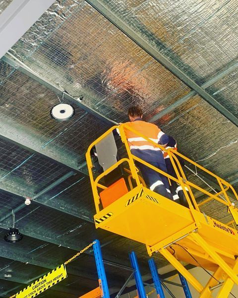 A Man Is Standing on A Yellow Lift Looking up At the Ceiling — Paton's Electrical Services in Pialba, QLD