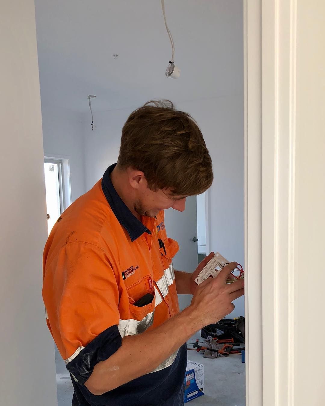 A Man in An Orange Shirt Is Working on A Light Switch — Paton's Electrical Services in Pialba, QLD