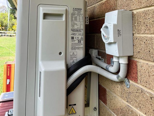 A White Air Conditioner Is Attached to A Brick Wall — Paton's Electrical Services in Hervey Bay, QLD