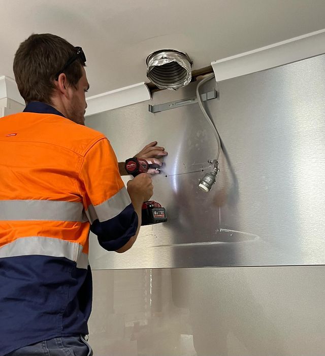 A Man Is Working on A Stainless Steel Wall with A Drill — Paton's Electrical Services in Urangan, QLD