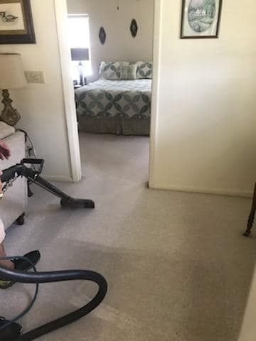 A person uses a vacuum cleaner on a beige carpet in a room that opens into a bedroom with a patterned bedspread.