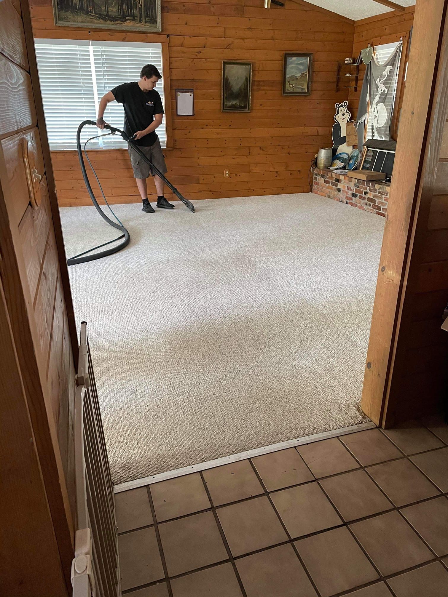 A person uses a professional carpet cleaning wand on a light-colored carpet inside a wood-paneled room.