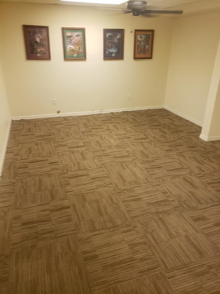 A room with tan walls, brown patterned carpet tiles, and four framed pictures hanging in a row on the back wall.