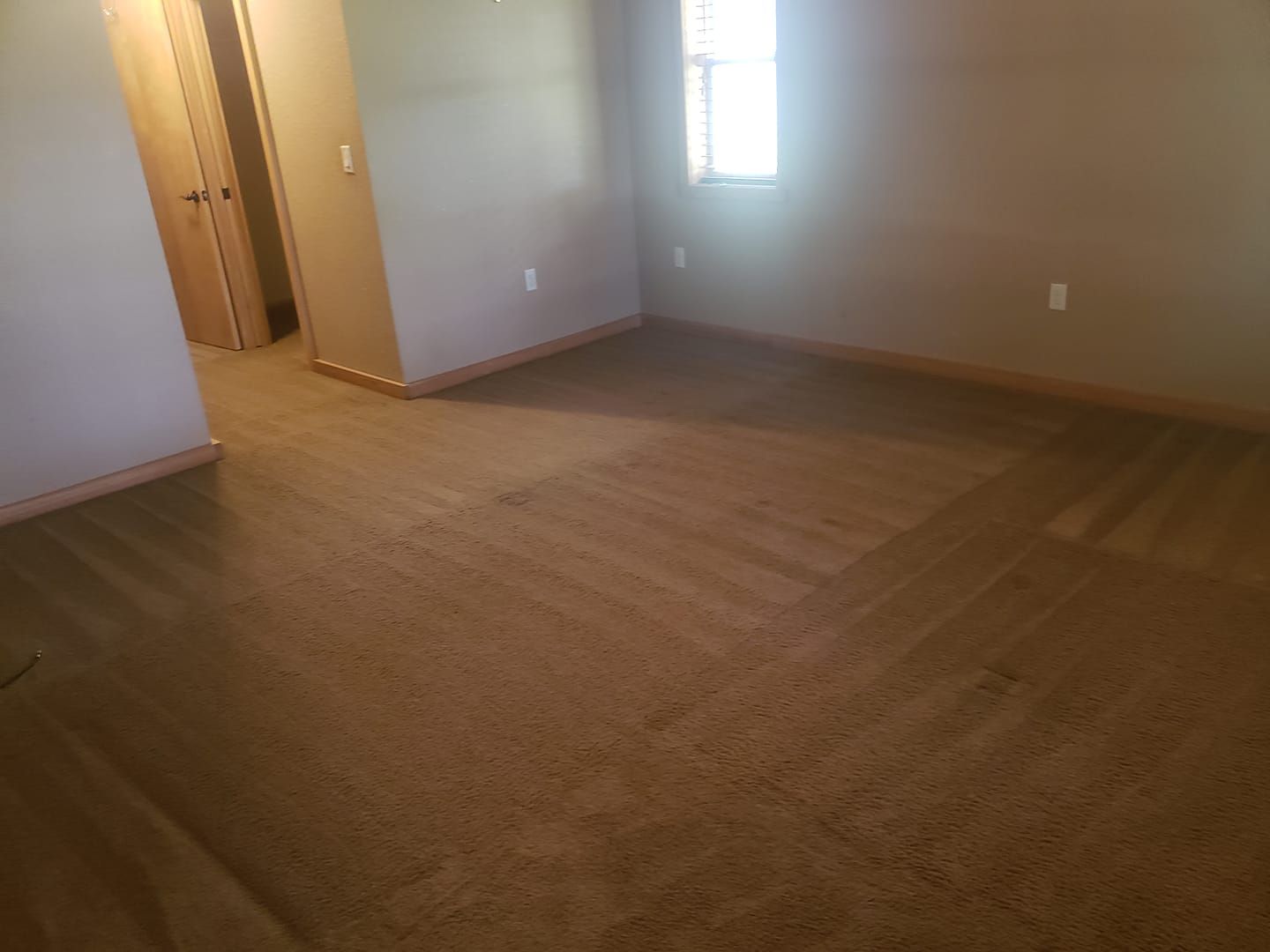 An empty room with beige carpet, light brown walls, a window on the far wall, and a doorway on the left.