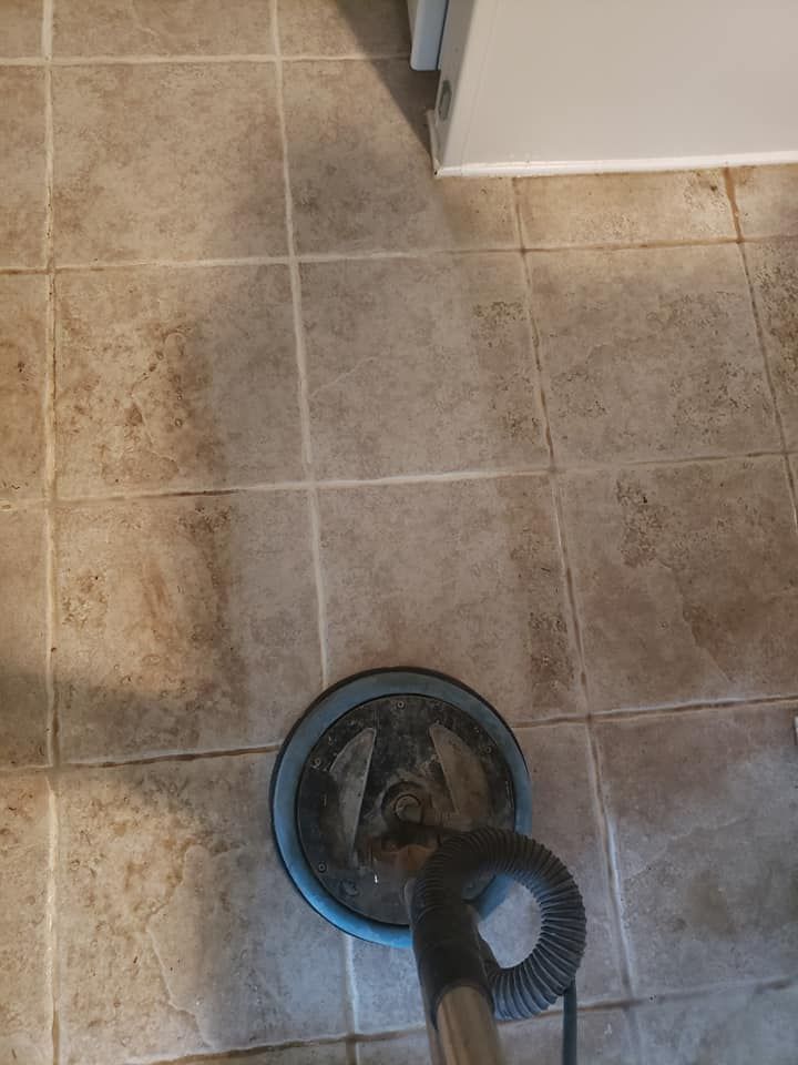 A floor cleaning machine scrubbing a beige, grid-patterned tiled floor, showing a clear contrast between dirty and clean.