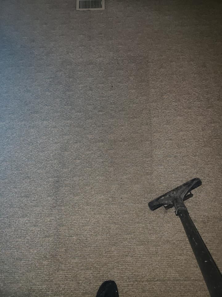 A vacuum cleaner floor attachment rests on a textured, light brown carpet in a room with a floor vent in the background.