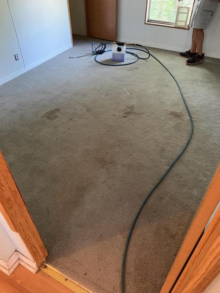 A person stands in a room with a gray carpeted floor, preparing to use carpet cleaning equipment connected by a hose.