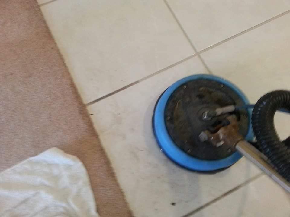 A blue floor cleaning machine sits on beige tiled flooring next to a carpeted area.