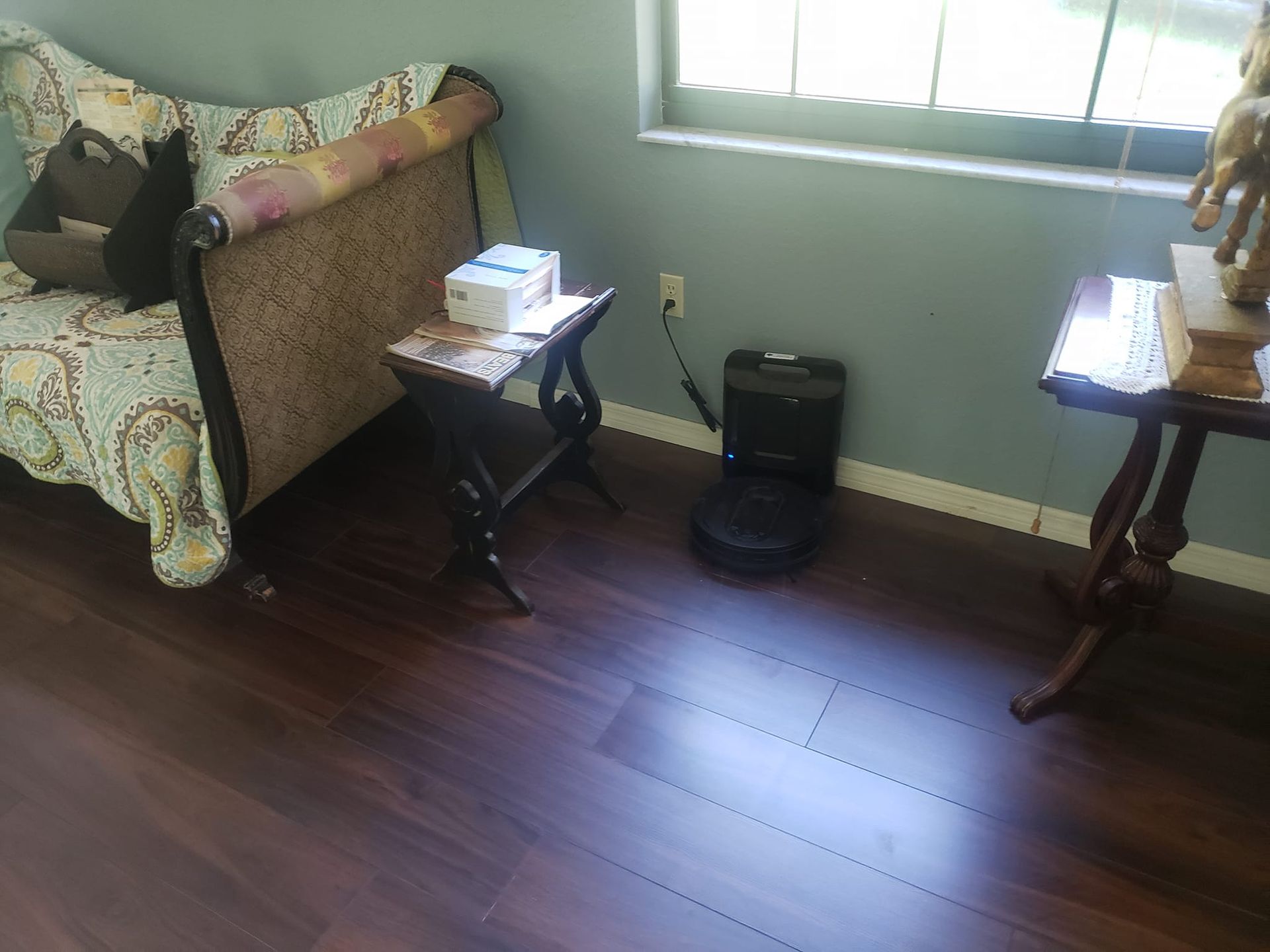 A robot vacuum sits on a wood floor between a patterned sofa, a small side table with a box, and a wooden pedestal table.